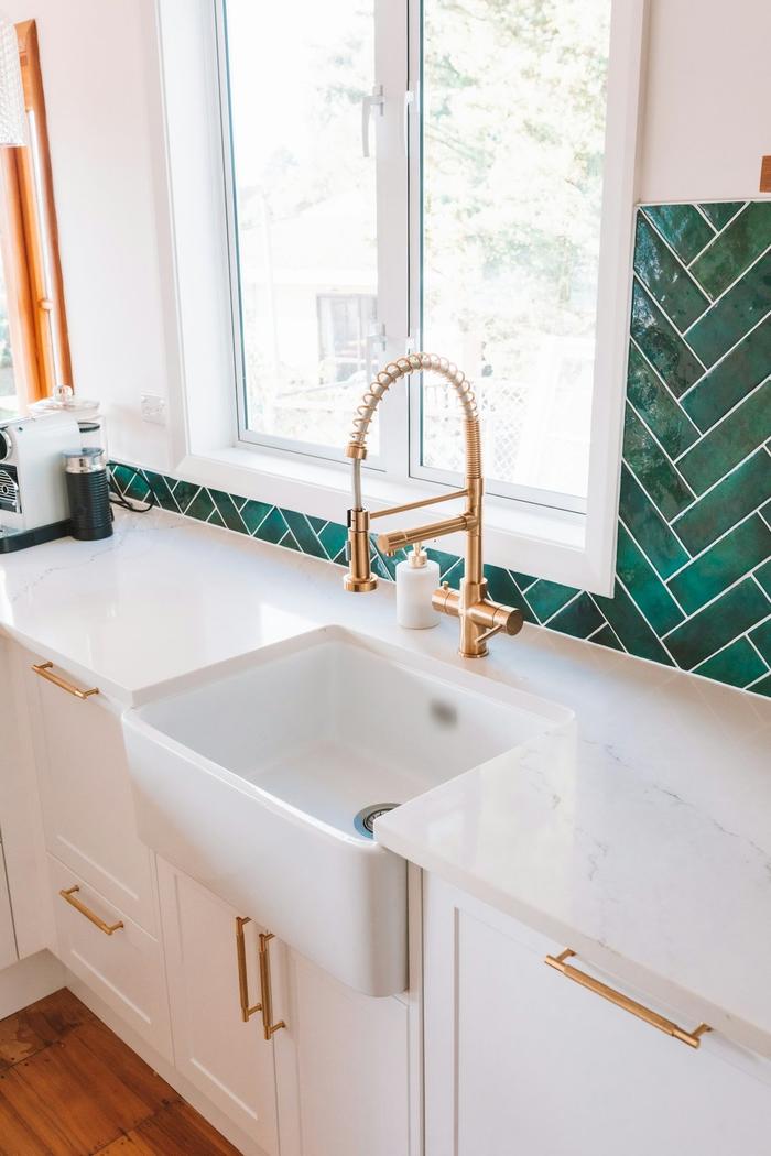 A white Belfast-style kitchen sink with a brass spring-neck tap, marble-effect worktop and green herringbone tile splashback.