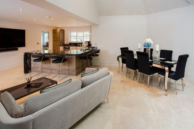 An open-plan kitchen, living and dining room with a grey sofa, a dark stone breakfast bar with stools and a black dining table on a pale tiled floor.