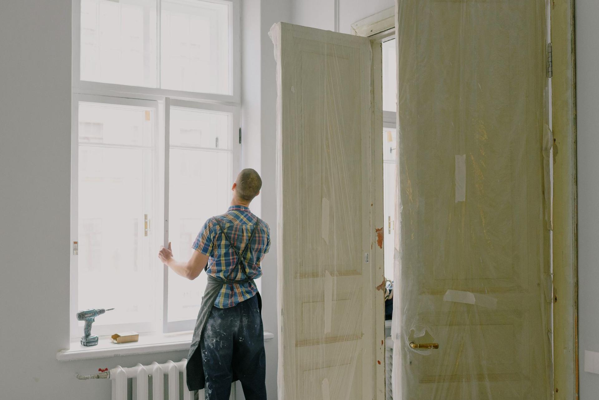 A tradesman in a checked shirt and work apron, seen from behind, fitting a tall window in a room under renovation.