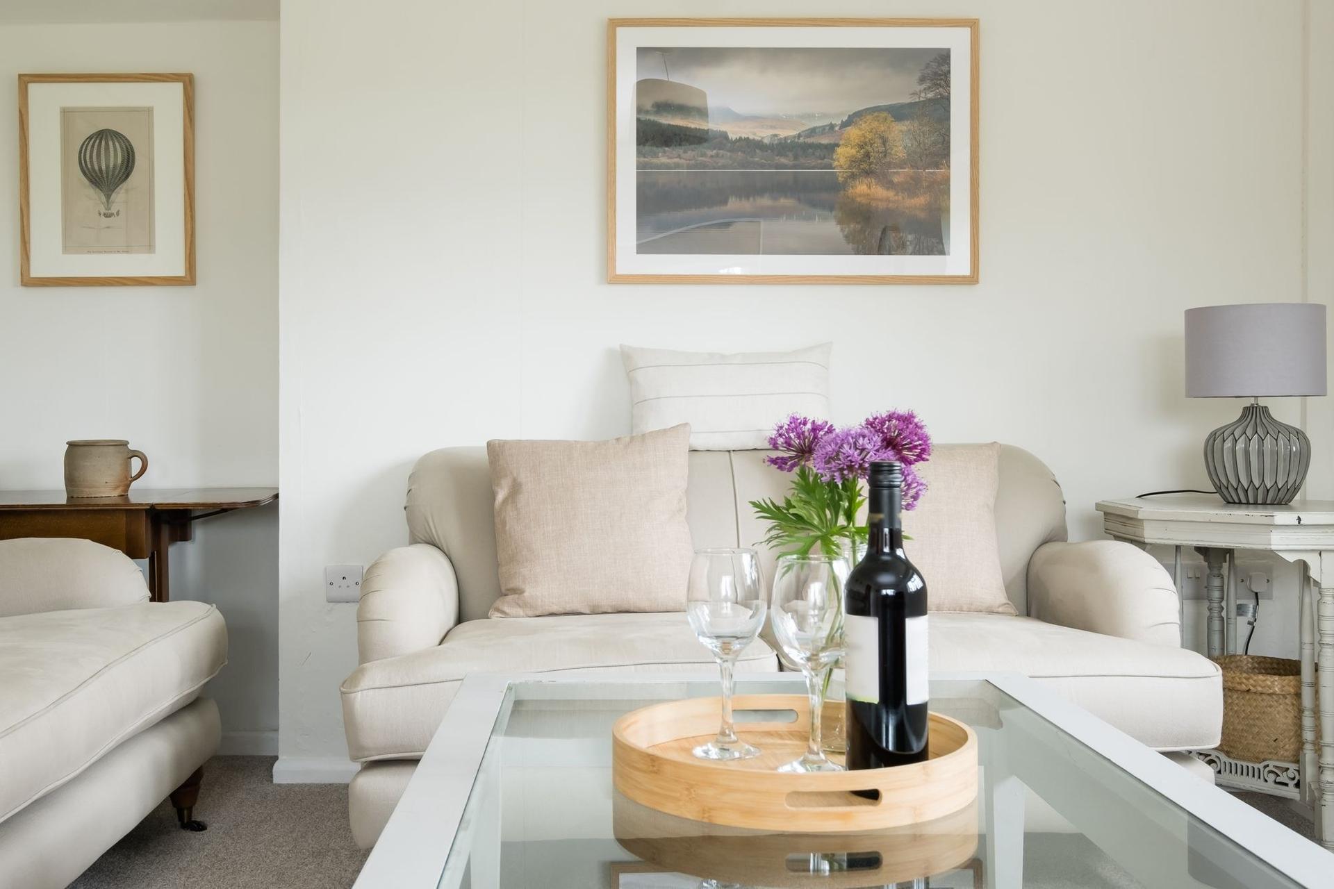 A bright cottage living room with cream sofas, framed prints on pale walls, a table lamp and a glass coffee table holding a tray of wine glasses and flowers.