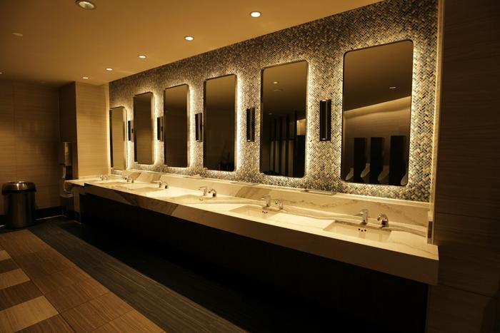 A warmly lit washroom with a long marble counter of undermount basins and sensor taps beneath backlit mirrors on a mosaic-tiled wall.
