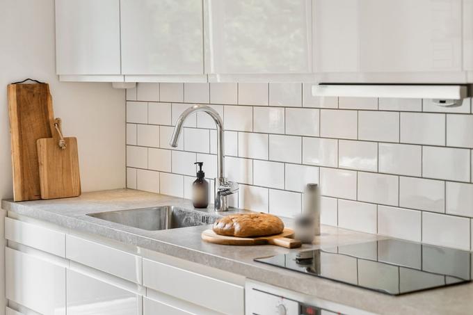 A white kitchen with a grey worktop, inset stainless steel sink, white metro-tile splashback and a loaf of bread on a board.