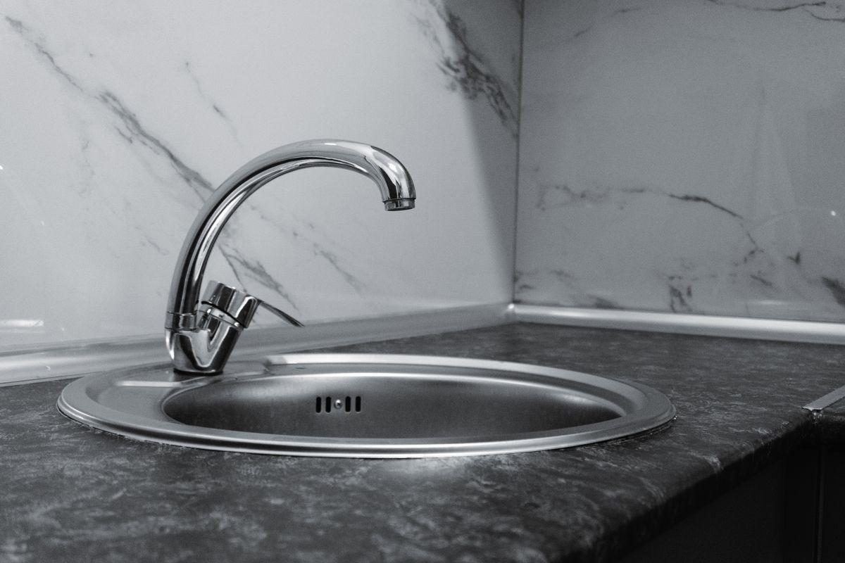 Black-and-white close-up of a round stainless steel sink and chrome tap set into a dark worktop against a marble-effect splashback.