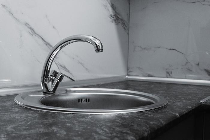 Black-and-white close-up of a round stainless steel sink and chrome tap set into a dark worktop against a marble-effect splashback.