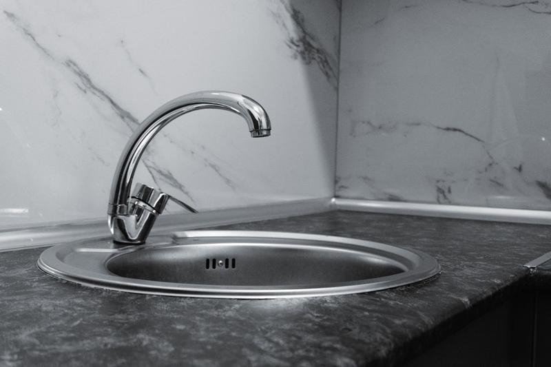 Black-and-white close-up of a round stainless steel sink and chrome tap set into a dark worktop against a marble-effect splashback.