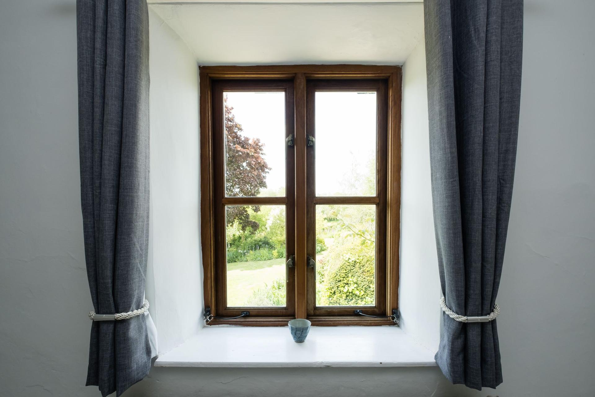 A timber casement window set in a deep white plastered reveal, with a white window board and grey curtains tied back on either side.