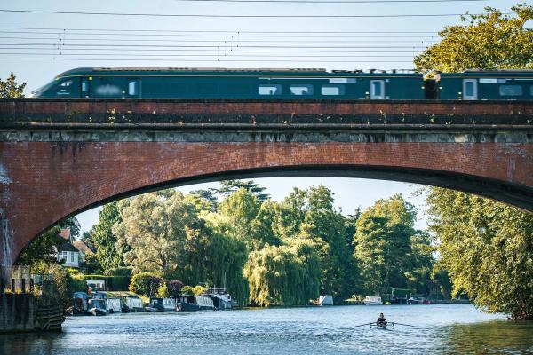 A train crossing Brunel's wide brick-arched railway bridge over the River Thames at Maidenhead, with narrowboats moored under willow trees.