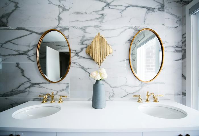 Twin undermount basins with brass taps in a white vanity top, below two oval gold-framed mirrors on marble-effect wall tiles.