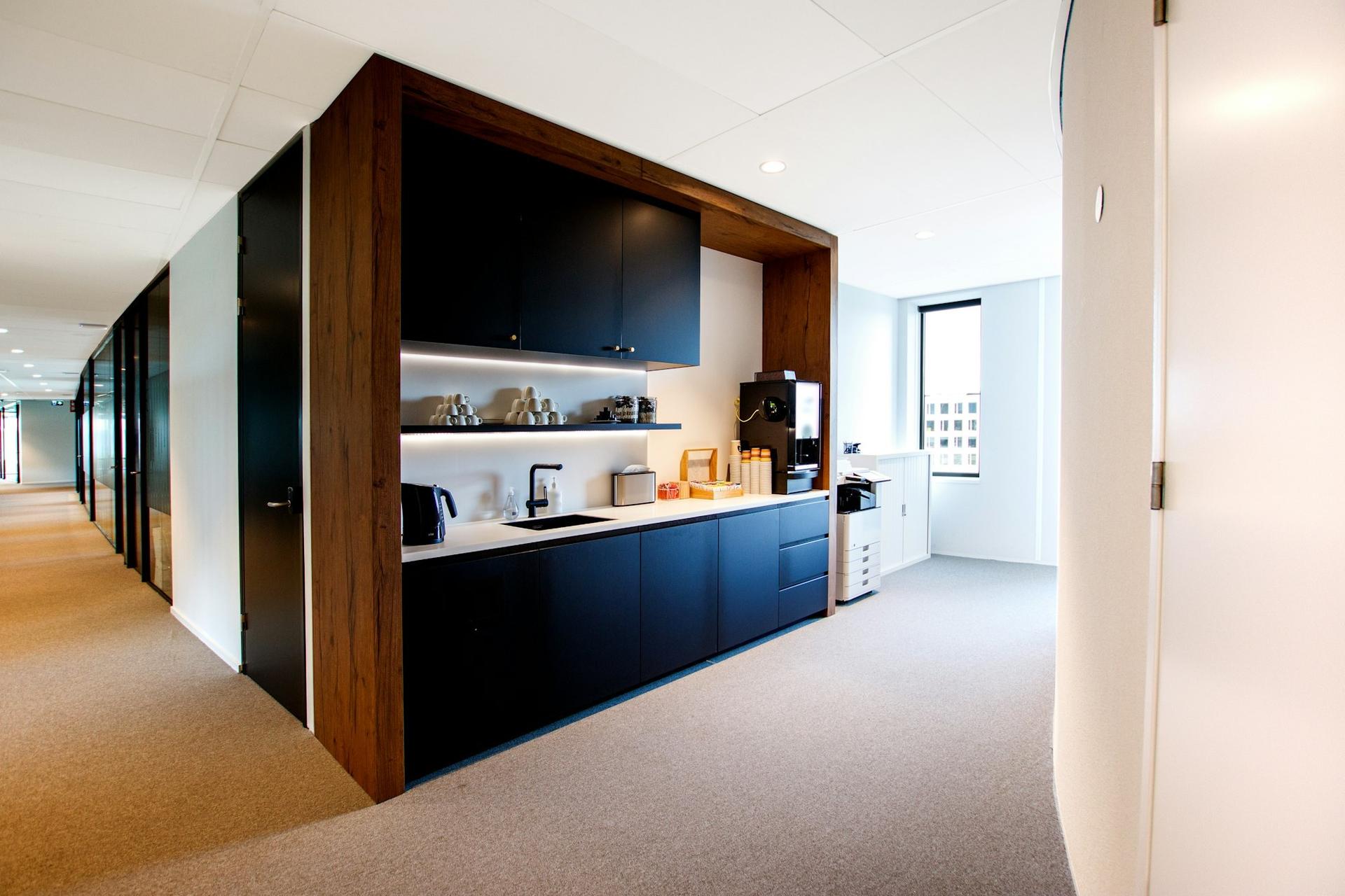 An office kitchenette with dark cabinets set in a timber surround, a white worktop and a coffee machine, beside a carpeted corridor.