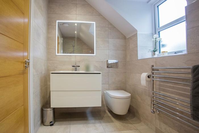 A newly fitted bathroom with a wall-hung toilet, white floating vanity unit, illuminated mirror and chrome towel rail under a sloping ceiling.