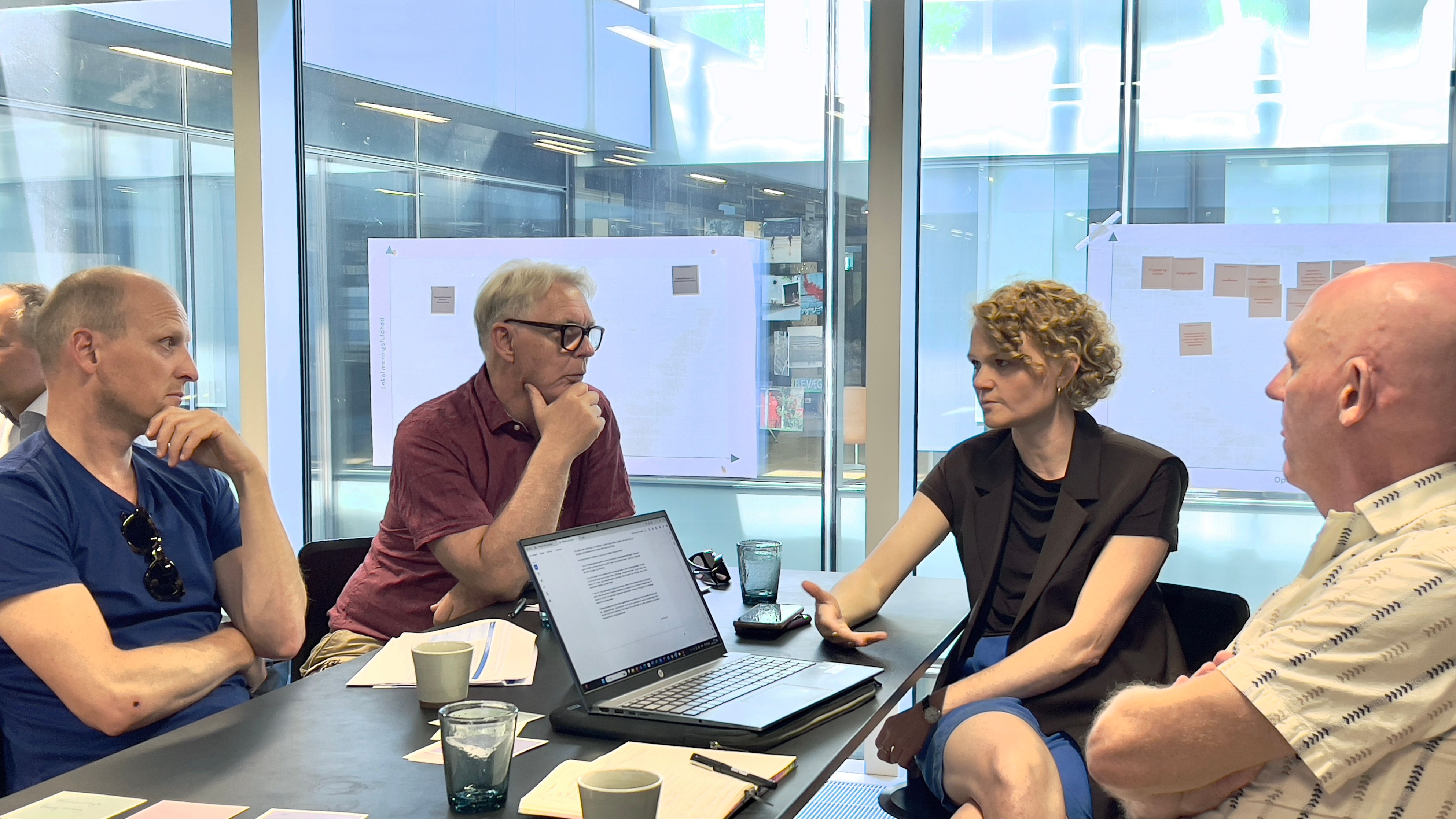 Fire mennesker til workshop i mødelokale med plancher på væggene