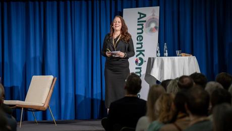 Ulla Jensen holder oplæg på scenen med AlmenKompas-logo i baggrunden