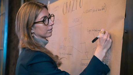 Kvinde med briller skriver med tus på planche, der hænger på en væg