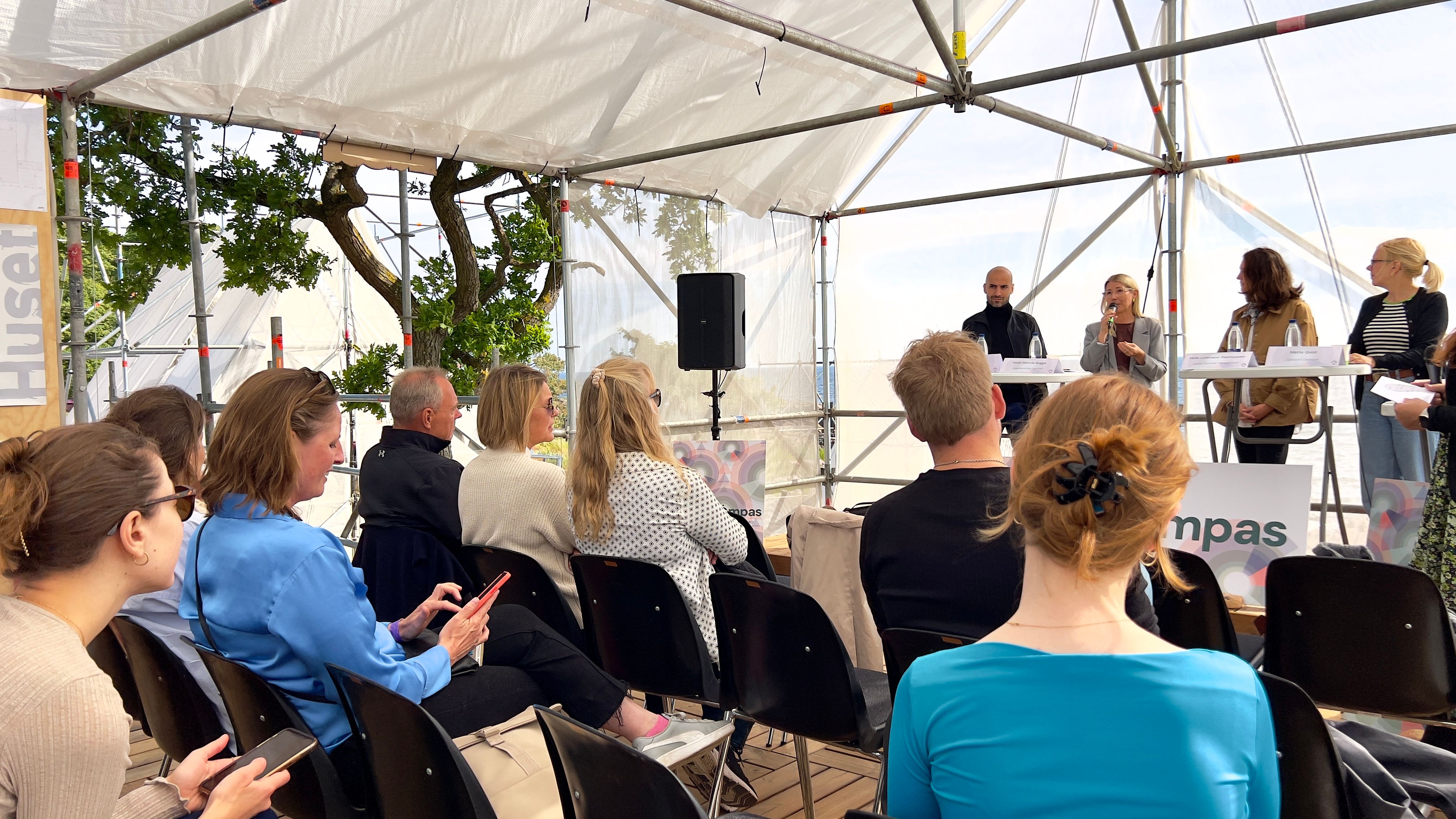 Paneldebat under gennemsigtig teltdug med fire kvinder i panelet. Forrest stolerækker med publikum.