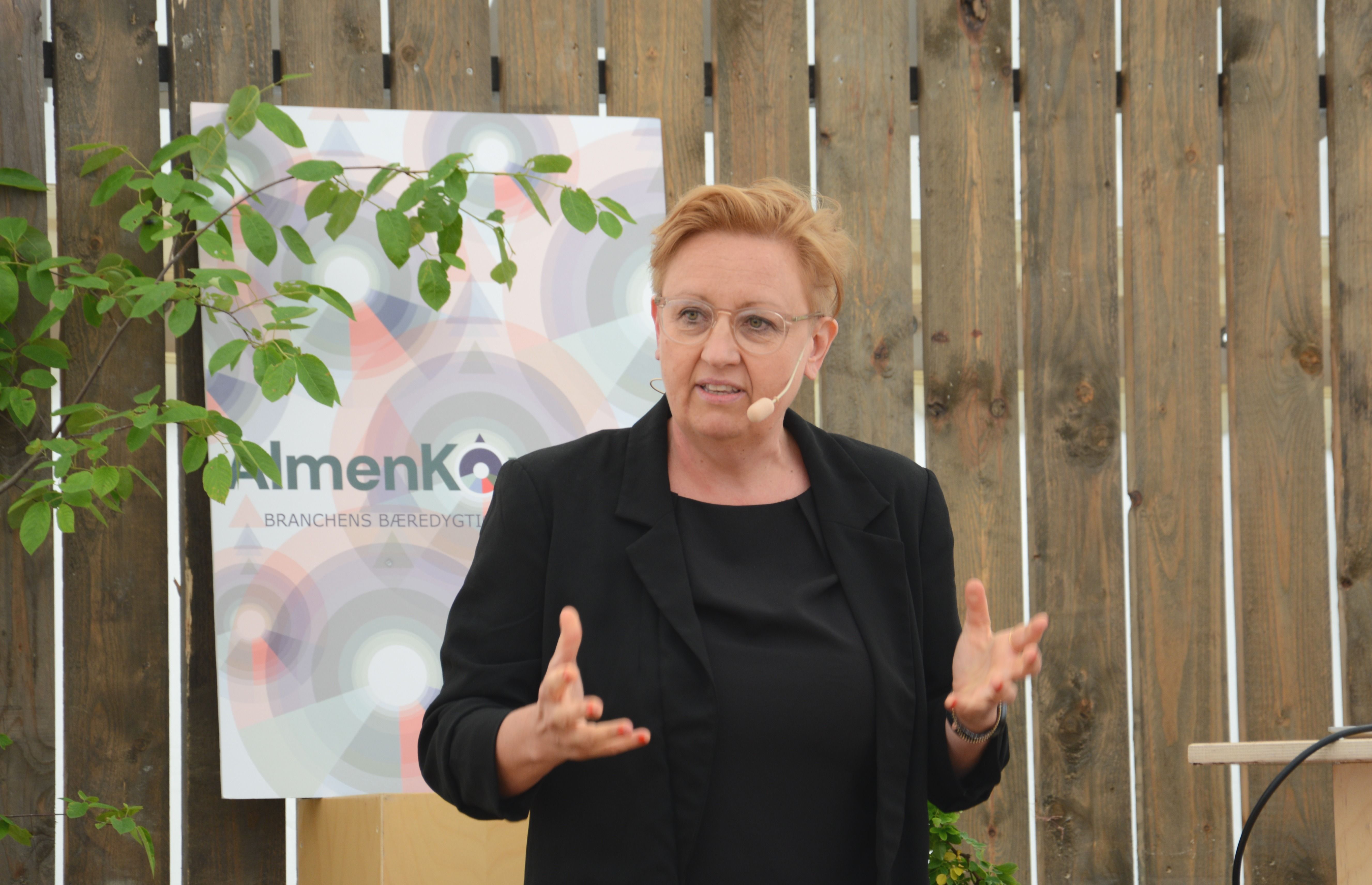 Kvinde holder oplæg på scene med bræddevæg og AlmenKompas-skilt i baggrunden
