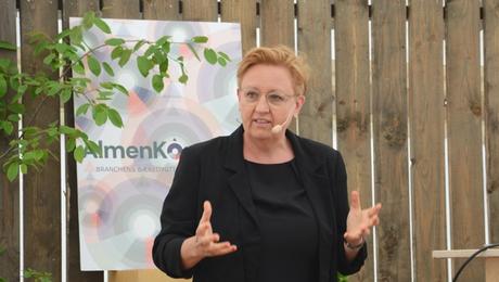 Kvinde holder oplæg på scene med bræddevæg og AlmenKompas-skilt i baggrunden