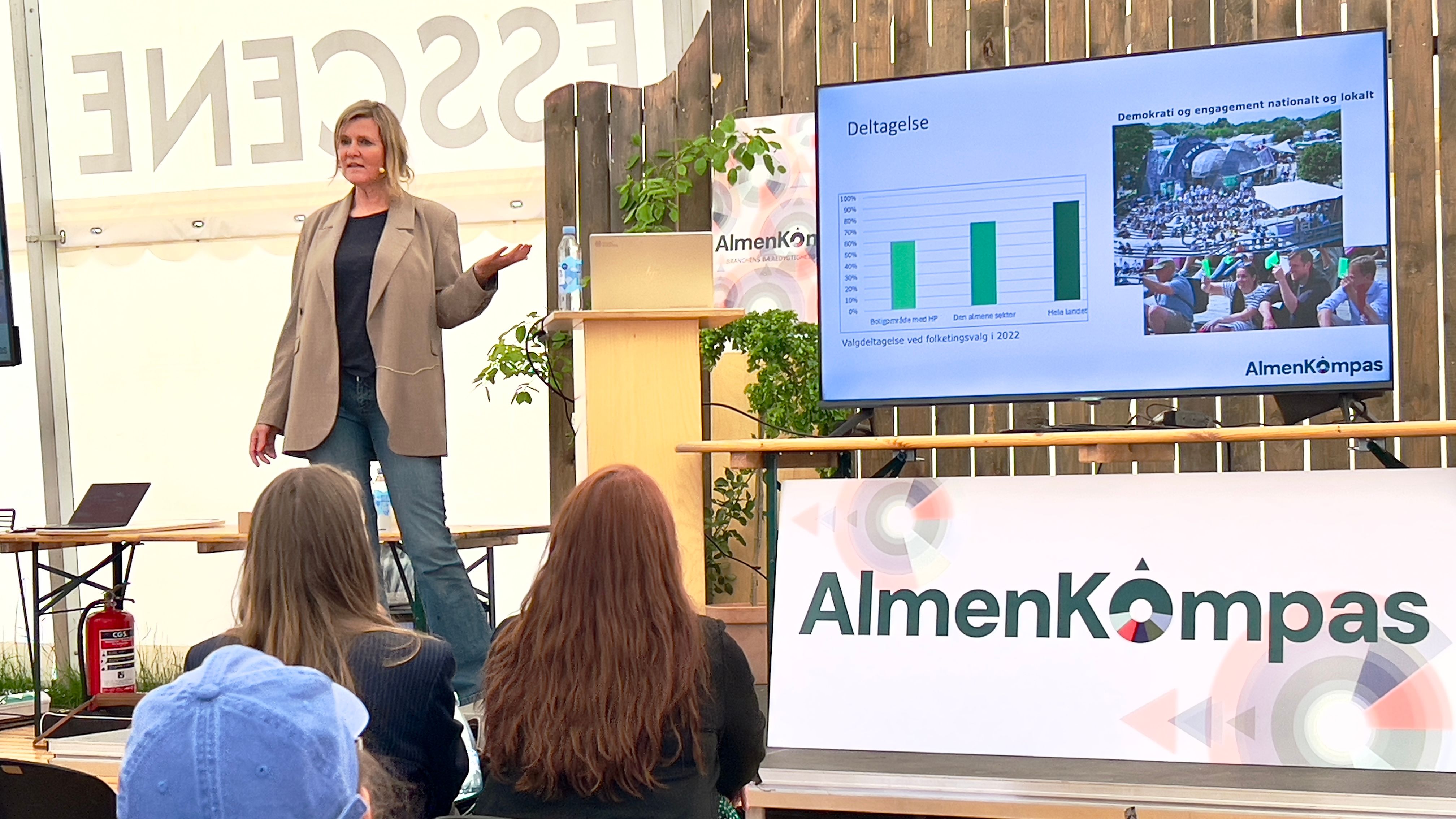 Kvinde på en scene holder oplæg og peger mod skærm med grafer. I forgrunden tilhørere.