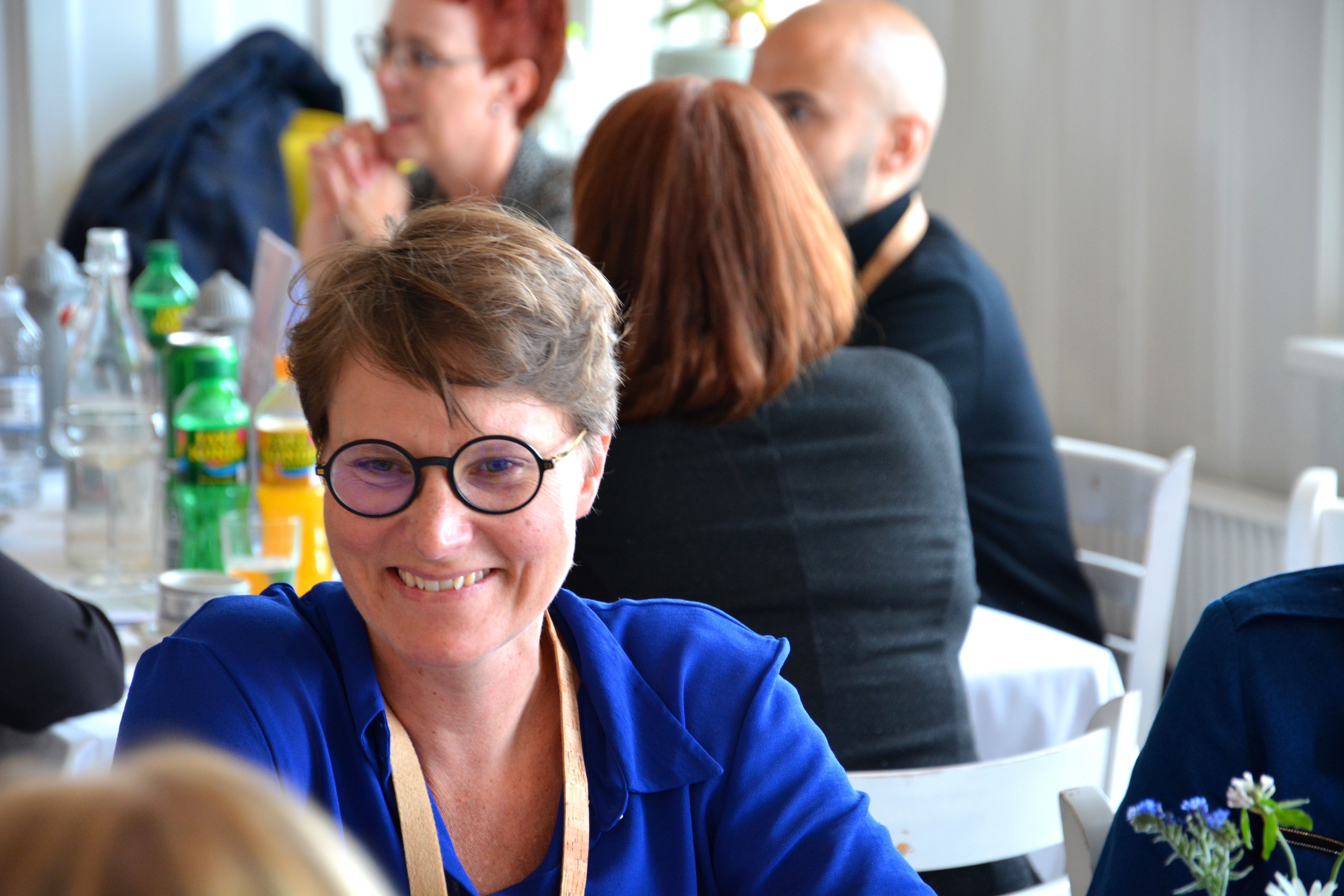 Kvinde med blå skjorte og briller sidder og smiler i restaurant med mange mennesker