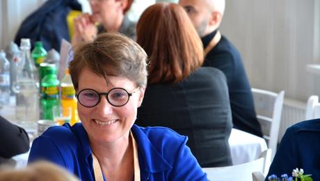 Kvinde med blå skjorte og briller sidder og smiler i restaurant med mange mennesker