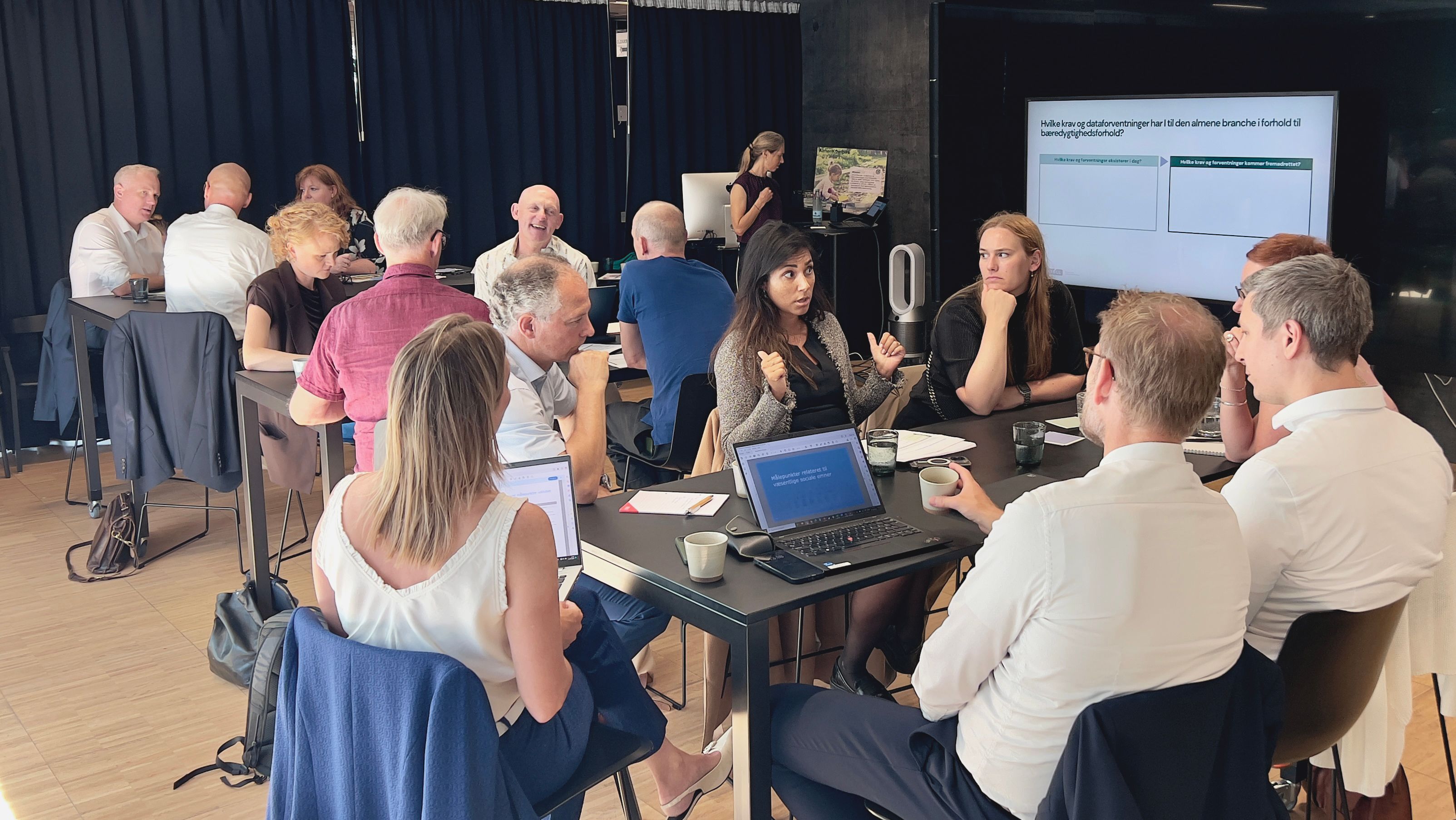 Cirka 15 mennesker sidder omkring tre borde og med skærm i baggrunden