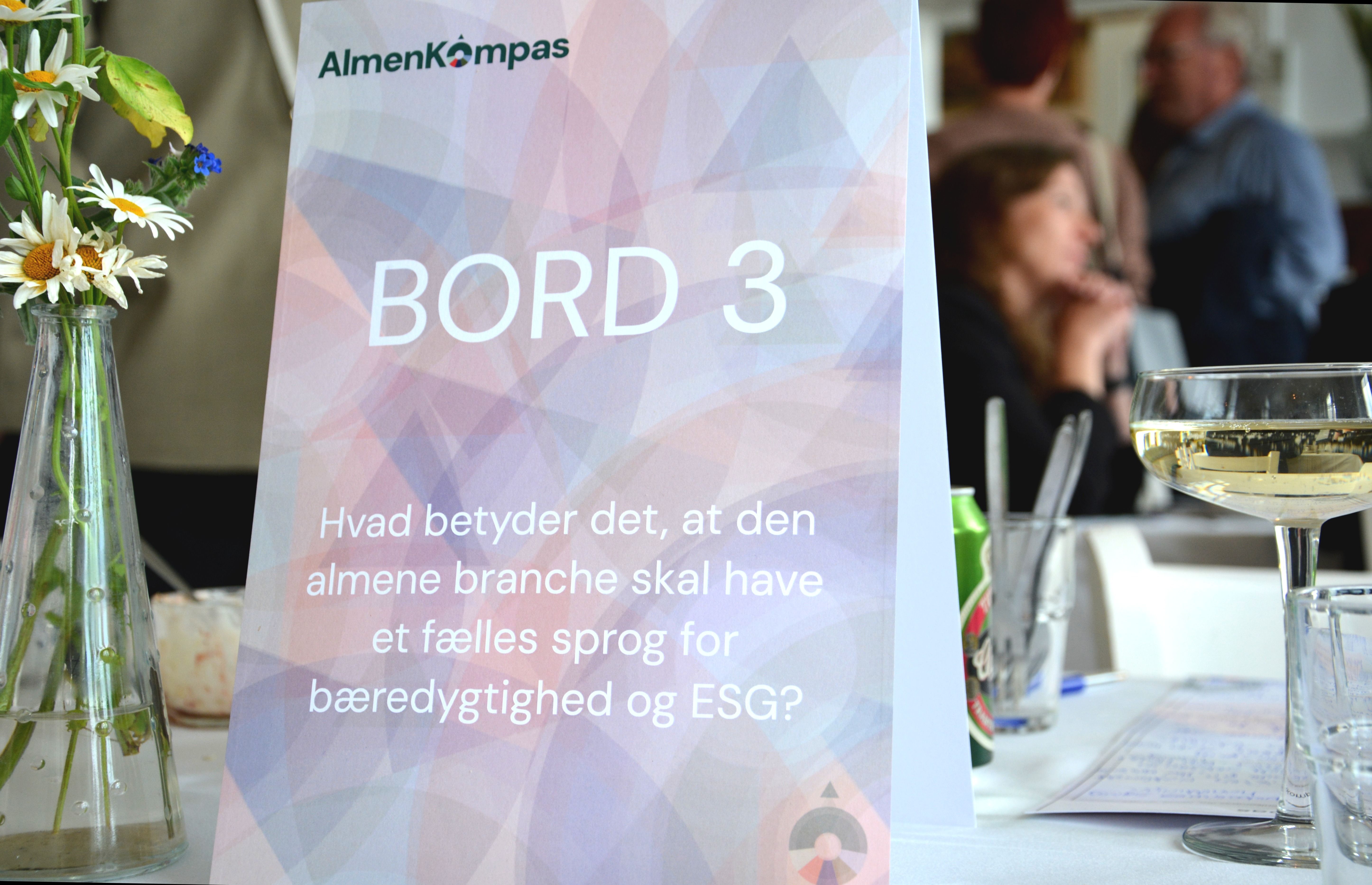 Skilt på bord med AlmenKompas-logo og mennesker i baggrund