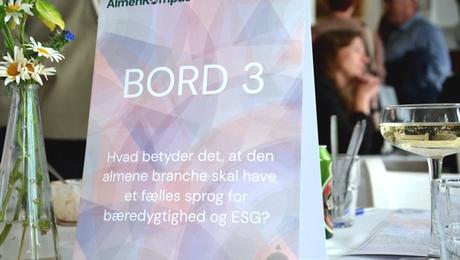 Skilt på bord med AlmenKompas-logo og mennesker i baggrund