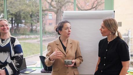 Tre kvinder i samtale stående foran en flip-over og med park i baggrunden uden for vinduet