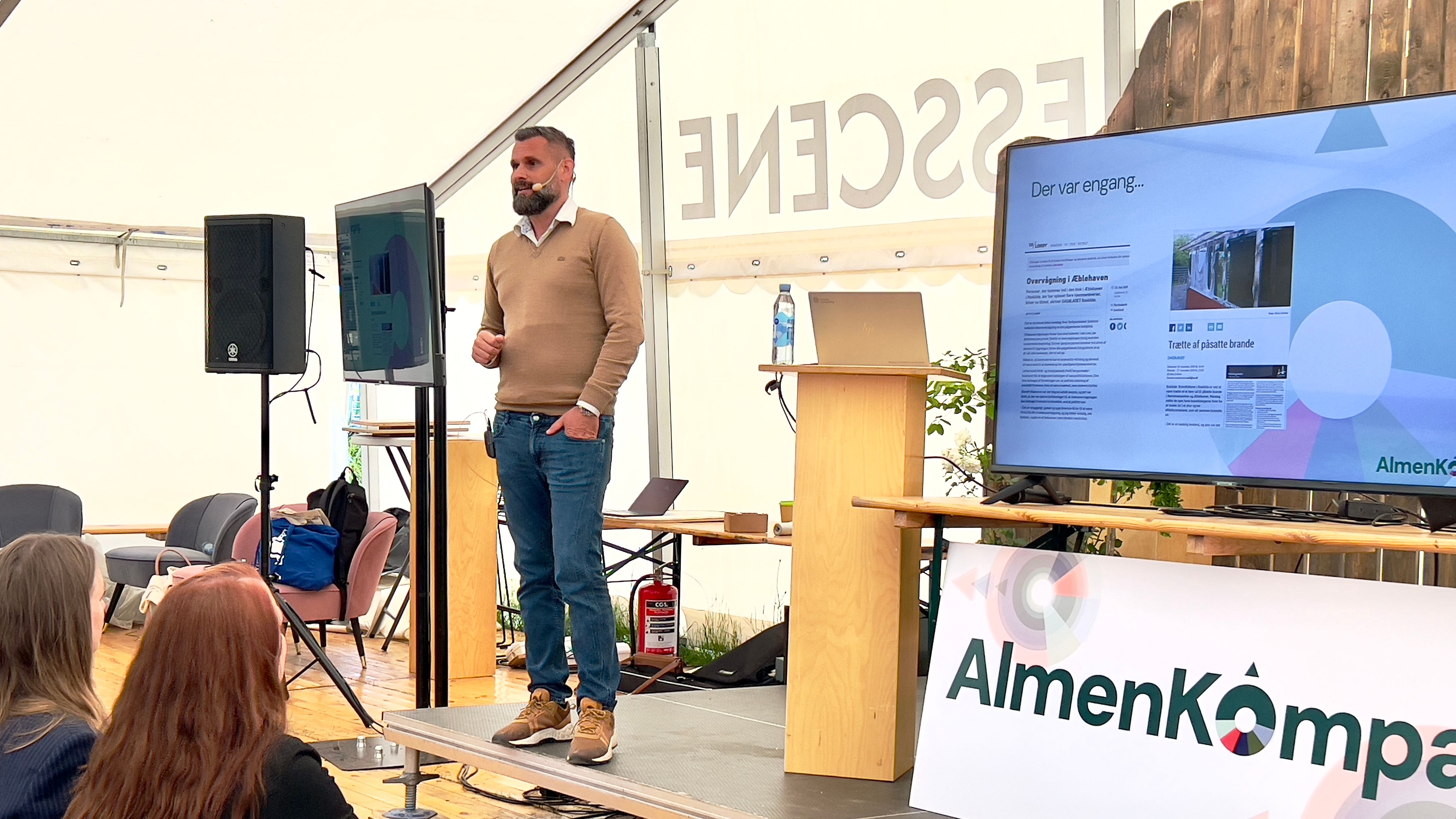 Mand med mikrofon på scene med AlmenKompas-logo i forgrund.