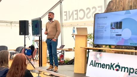 Mand med mikrofon på scene med AlmenKompas-logo i forgrund.