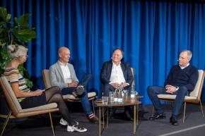 Paneldebat på scene med tre mænd og en kvinde i lave lænestole.
