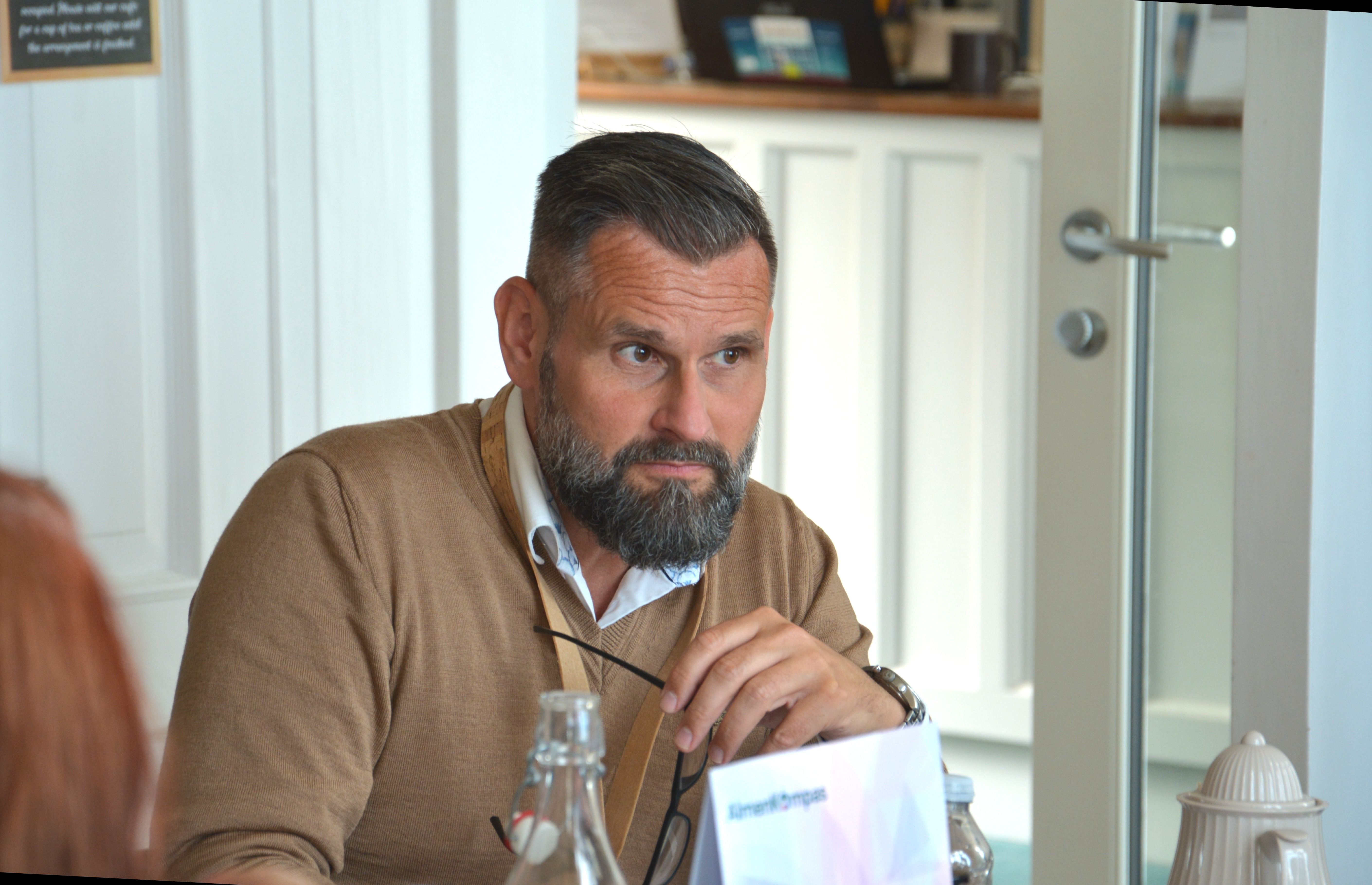 Mand med skæg i pullover og skjorte sidder ved bord og ser eftertænksom ud