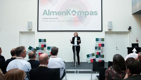 Connie Hedegaard på scenen foran publikum og med skærm med AlmenKompas-logo i baggrunden