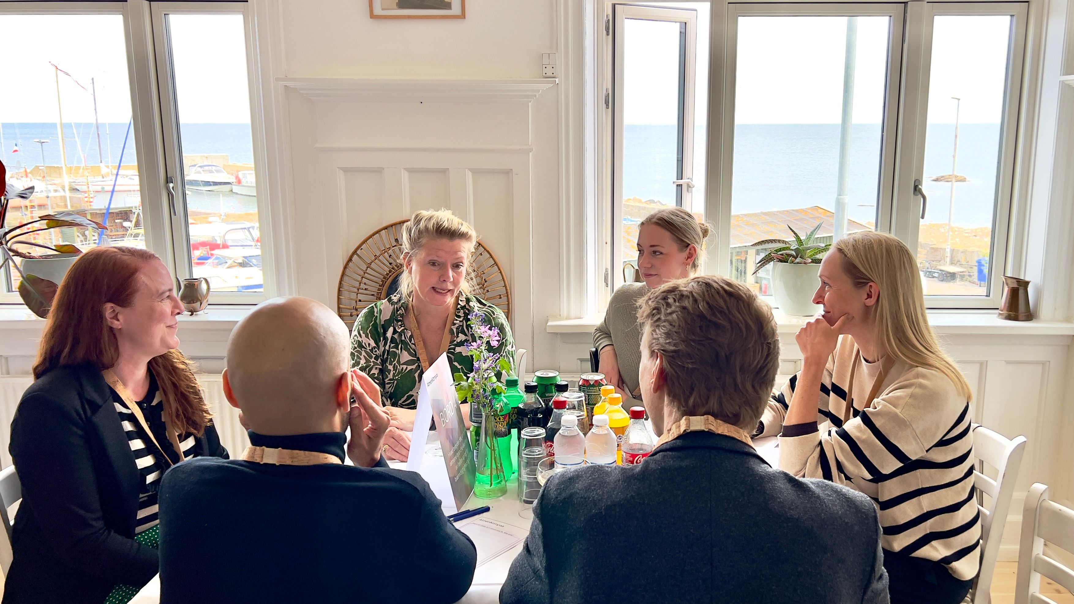 Fire kvinder og to mænd i samtale omkring bord med hvid dug og havn uden for vinduet i baggrunden