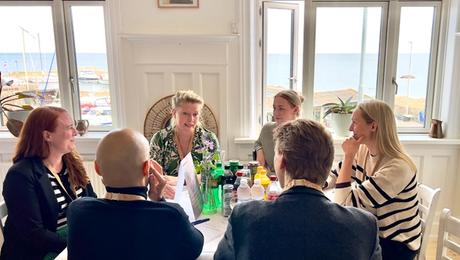 Fire kvinder og to mænd i samtale omkring bord med hvid dug og havn uden for vinduet i baggrunden