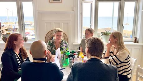 Fire kvinder og to mænd i samtale omkring bord med hvid dug og havn uden for vinduet i baggrunden