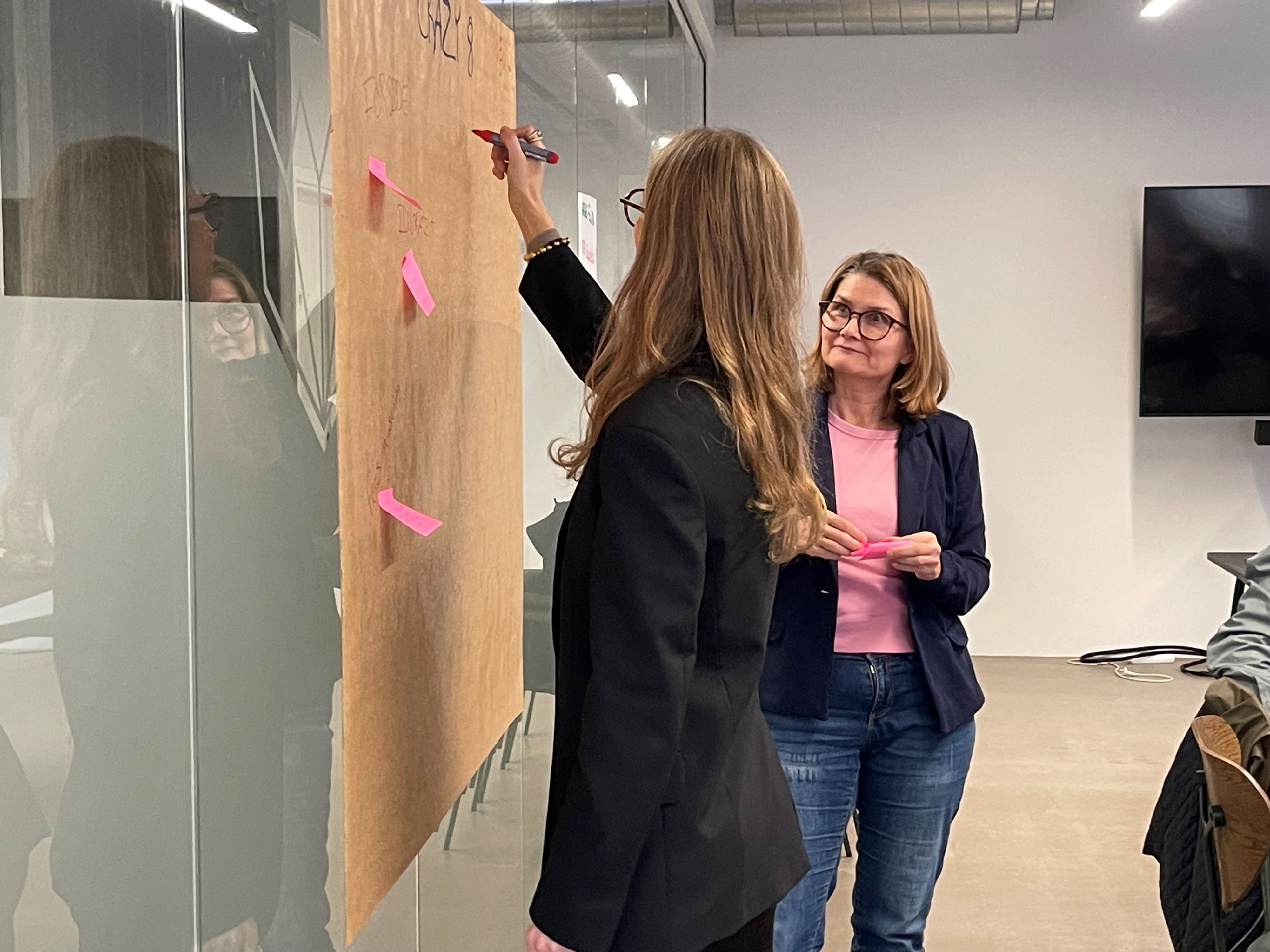 To kvinder står ved stor brun planche hængt op på en væg. Den ene noterer med tus