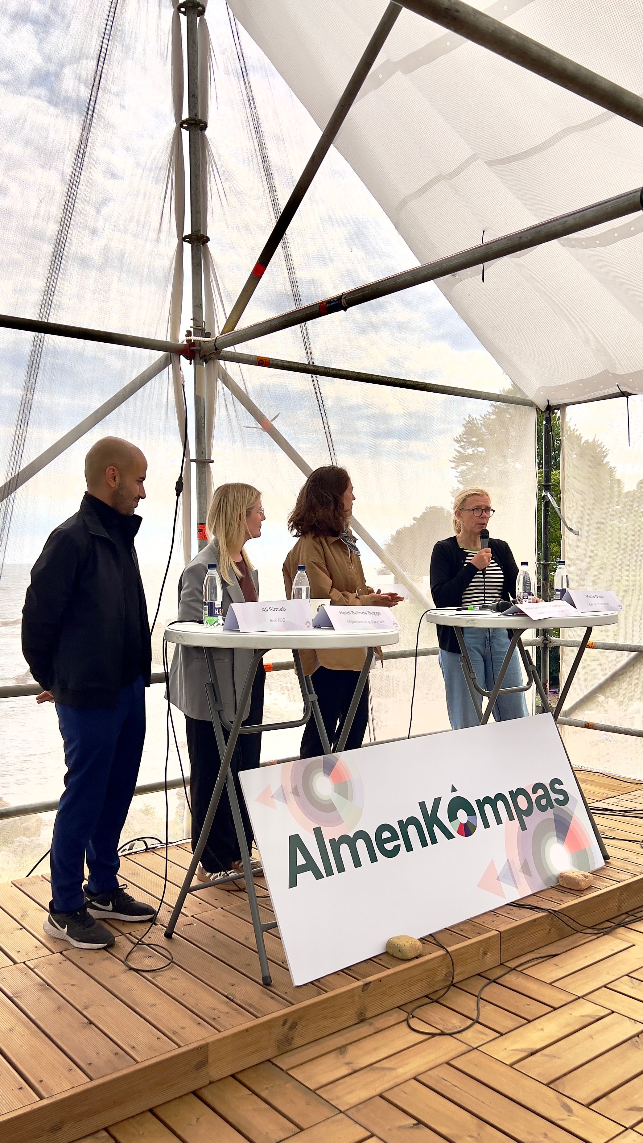 Tre kvinder og en mand står ved høje borde i paneldebat i telt.