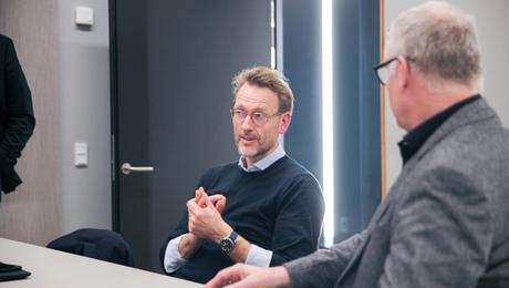 To mænd ved bord i mødelokale. En taler, mens den anden lytter