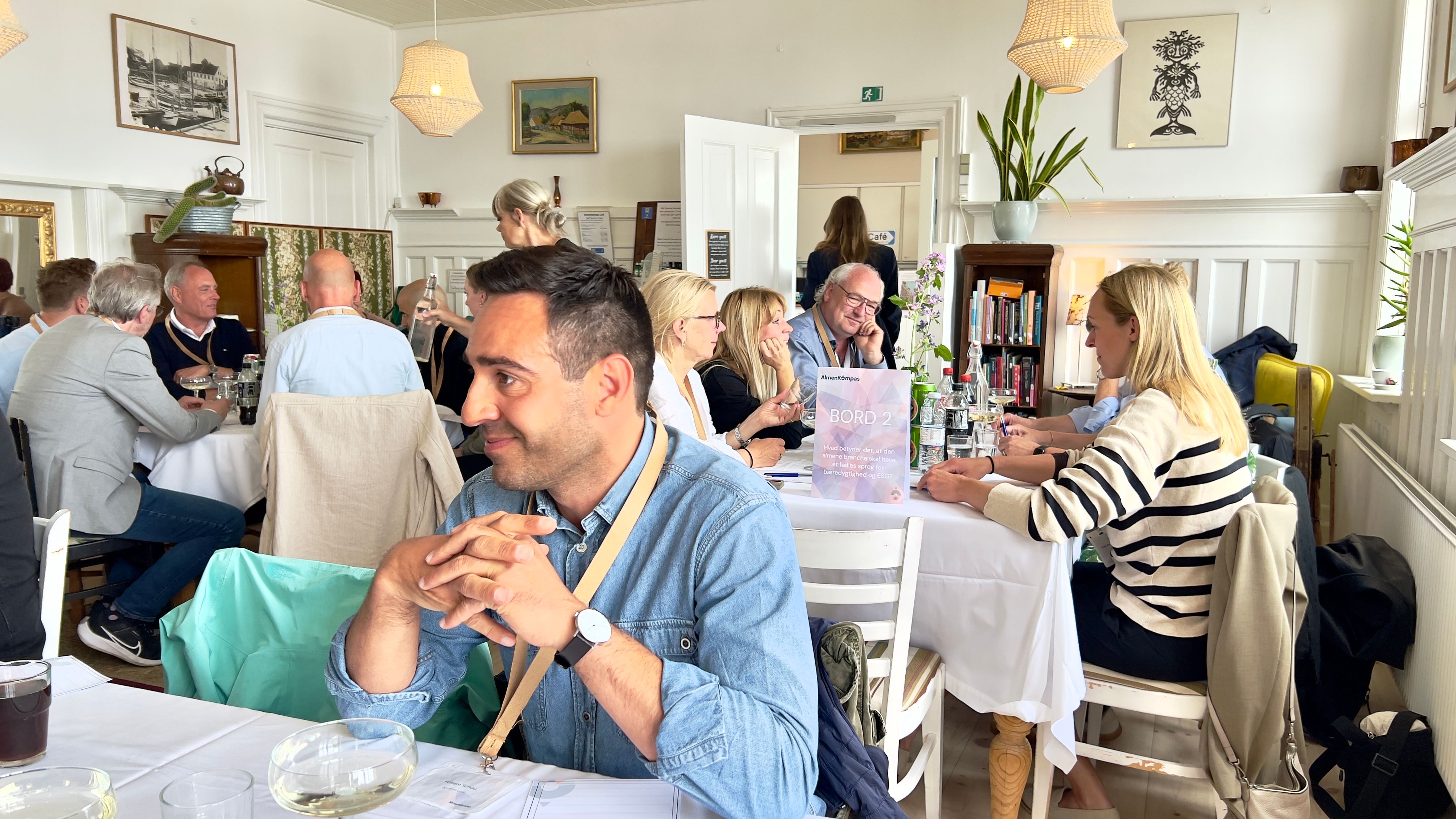 Mange mennesker ved borde i restaurant. Mand i blå skjorte i forgrunden.