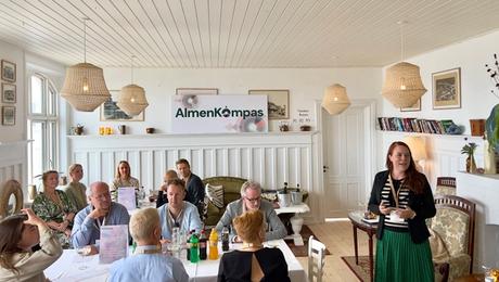 Restaurant med ca. 10 mennesker omkring borde med hvide duge samt kvinde der står og taler