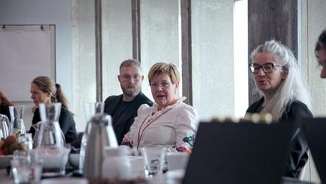 Fem personer siddende ved bord i mødelokale