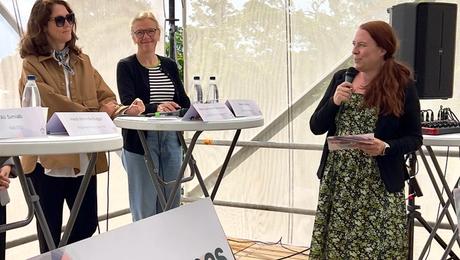 Paneldebat i telt med trægulv. To kvinder i panelet og en kvindelig moderator med mikrofon
