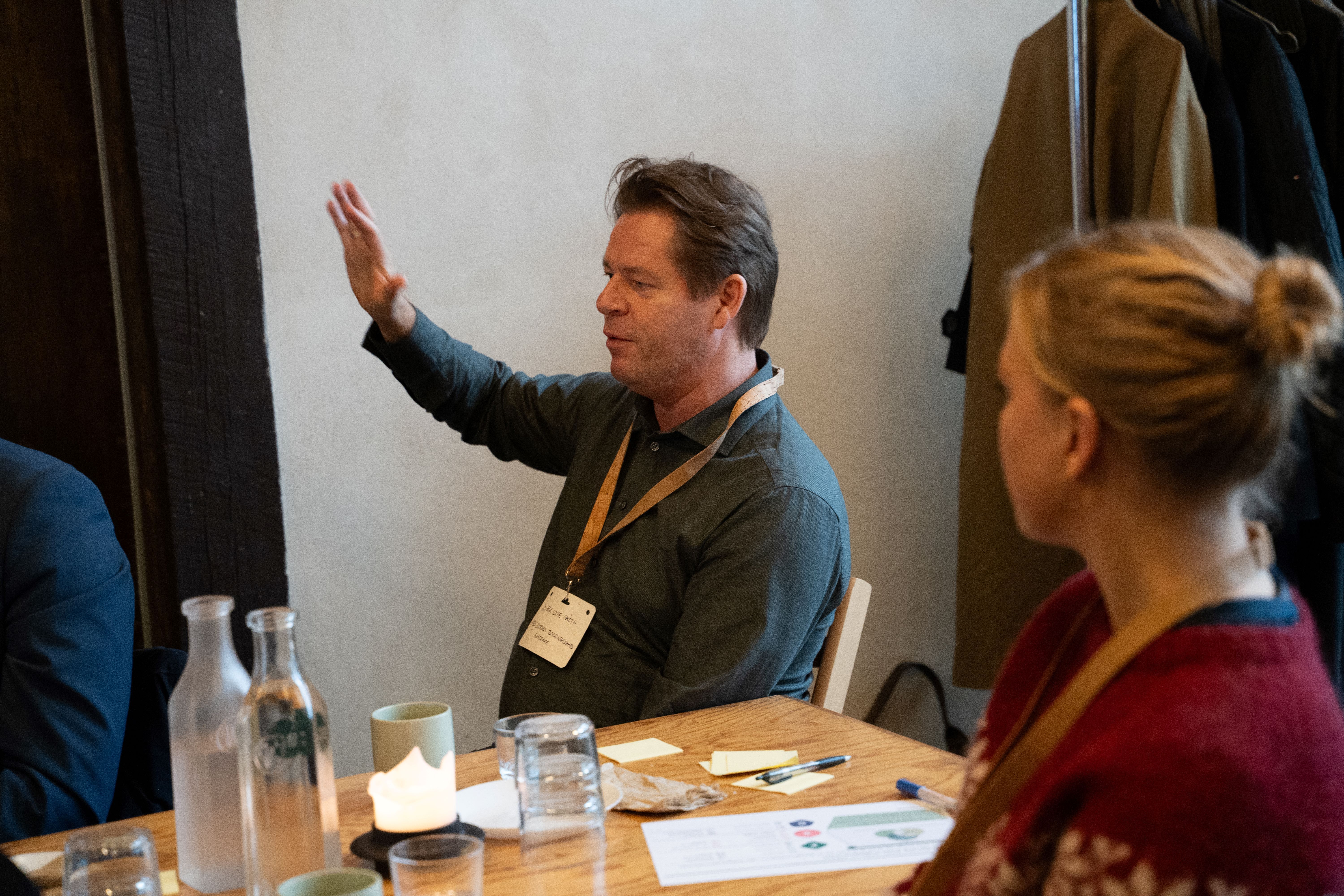 Mand sidder ved bord og gestikulerer med højre hånd, mens en kvinde kigger og lytter