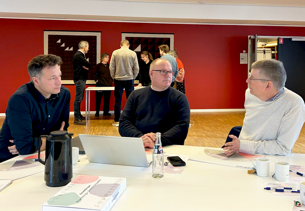 To mandlige deltagere til læringsdag ved bord - gruppe af mennesker i baggrunden