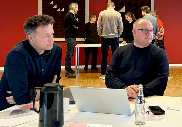 To mandlige deltagere til læringsdag ved bord - gruppe af mennesker i baggrunden