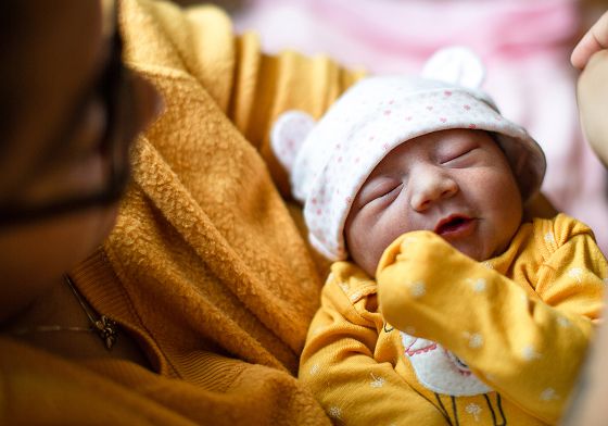 Close-up of a newborn baby in a yellow outfit and bear hat being held gently by an adult in a cozy indoor setting.