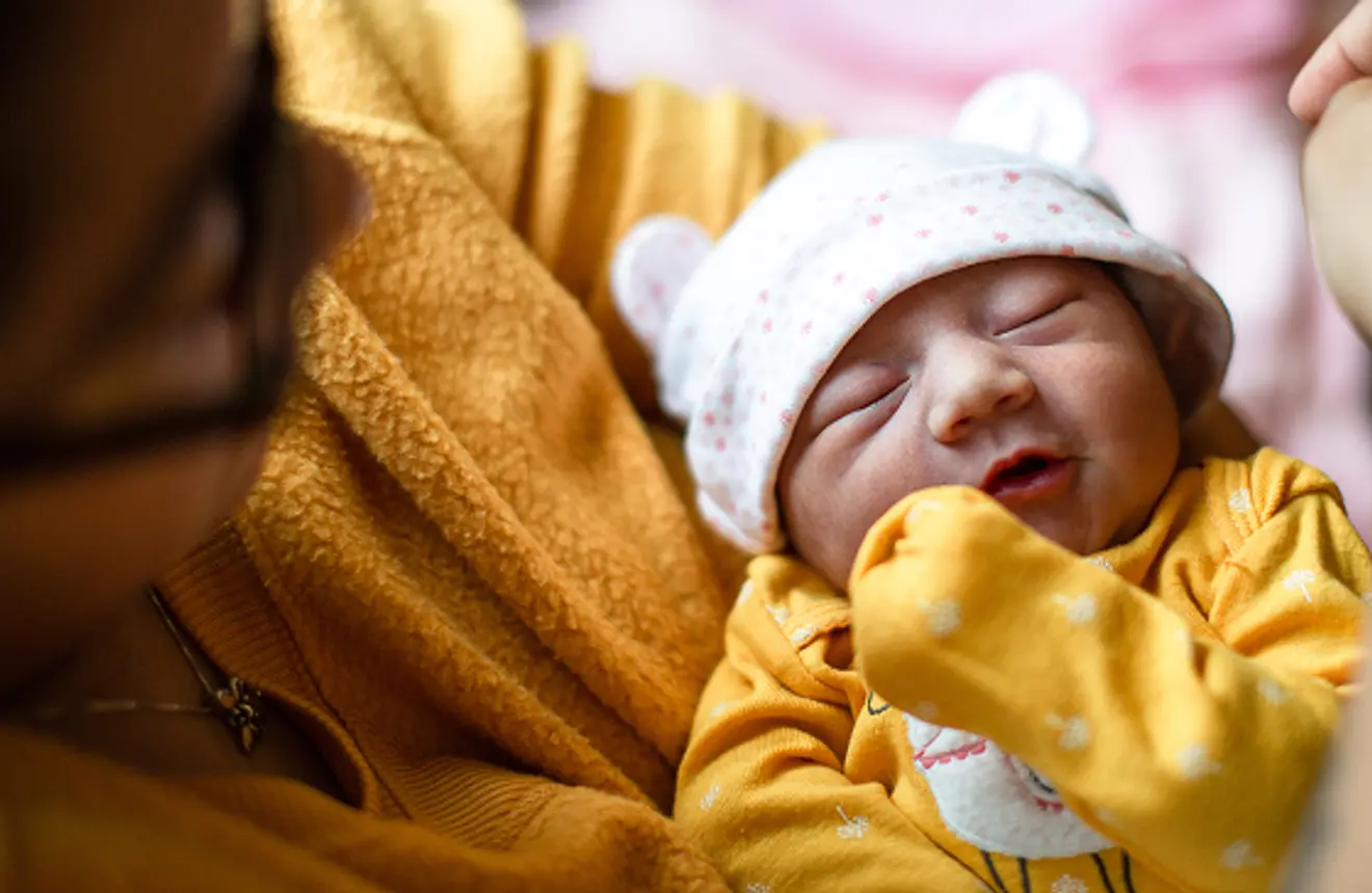 Close-up of a newborn baby in a yellow outfit and bear hat being held gently by an adult in a cozy indoor setting.