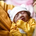 Close-up of a newborn baby in a yellow outfit and bear hat being held gently by an adult in a cozy indoor setting.