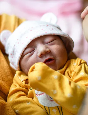 Close-up of a newborn baby in a yellow outfit and bear hat being held gently by an adult in a cozy indoor setting.
