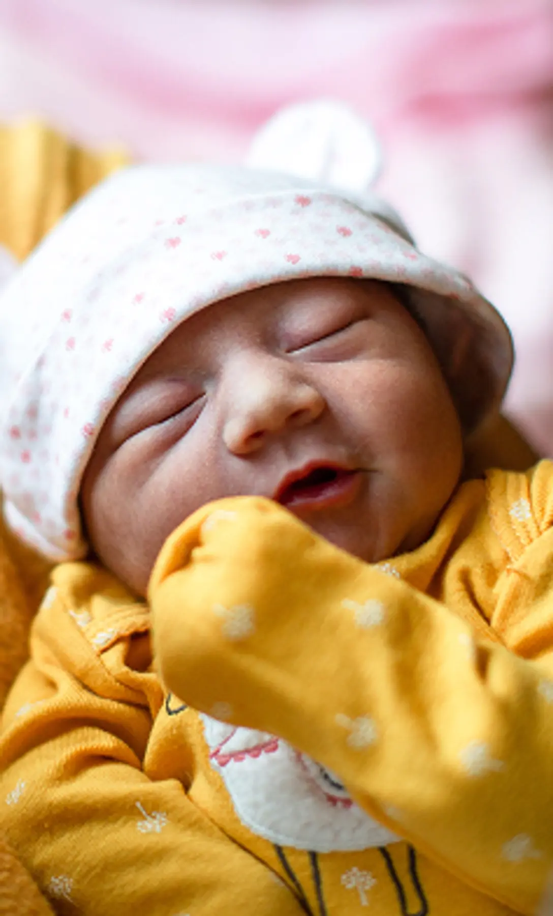 Close-up of a newborn baby in a yellow outfit and bear hat being held gently by an adult in a cozy indoor setting.